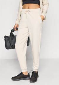 Μπεζ αθλητικό παντελόνι με ελαστική μέση και κορδόνι. Διαθέτει πλαϊνές τσέπες και στενά μπατζάκια. Συνδυάζεται με μαύρα αθλητικά παπούτσια και μαύρη τσάντα.