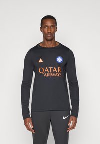 Μαύρο πουκάμισο με μακρύ μανίκι και λογότυπο "QATAR AIRWAYS" σε πορτοκαλί χρώμα, σε συνδυασμό με σκούρες γκρι παντελόνες που φέρουν λογότυπο Nike σε λευκό χρώμα.