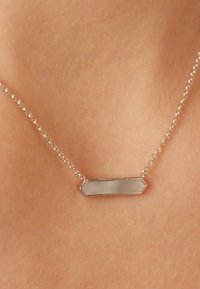 Zilveren ketting met een hexagonaal hangertje met een gladde, reflecterende oppervlakte, hangend tegen een huidskleurige achtergrond.