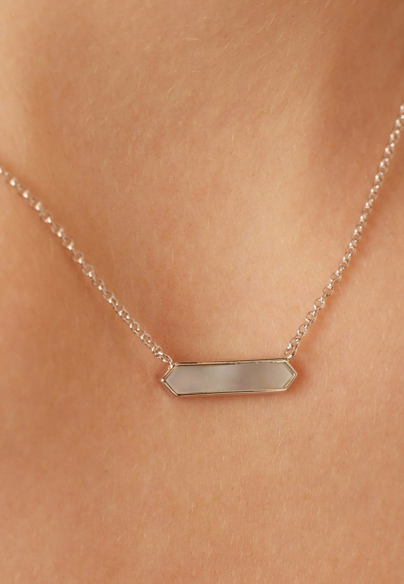 Zilveren ketting met een hexagonaal hangertje met een gladde, reflecterende oppervlakte, hangend tegen een huidskleurige achtergrond.