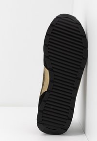 Semelle en caoutchouc noir avec un motif de bande de roulement strié sous une chaussure dorée et noire posée contre des murs blancs.