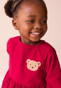 Vestito di velluto a coste rosso con maniche corte, caratterizzato da una toppa a forma di orsetto marrone chiaro sul petto. Tessuto liscio e gonna arricciata.