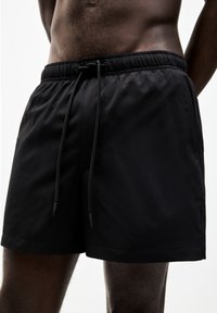 Schwarze Badeshorts aus strukturiertem Stoff mit einem elastischen Bund und Kordelzug. Verfügt über einen geraden Saum und dezente Steppdetails.