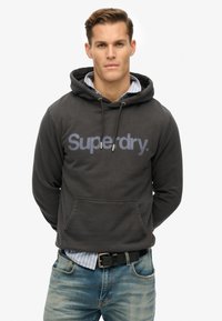 Sudadera negra de mezcla de algodón, con un gran logo de "Superdry." en la parte frontal, cordones de ajuste, bolsillo delantero y puños acanalados. Llevada sobre una camisa a rayas.