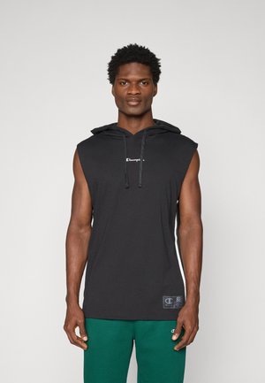 Muškarac u crnoj bezrukavnoj majici s kapuljačom i vezicama, te zelenim hlačama, stoji na jednobojnoj svijetloj pozadini.