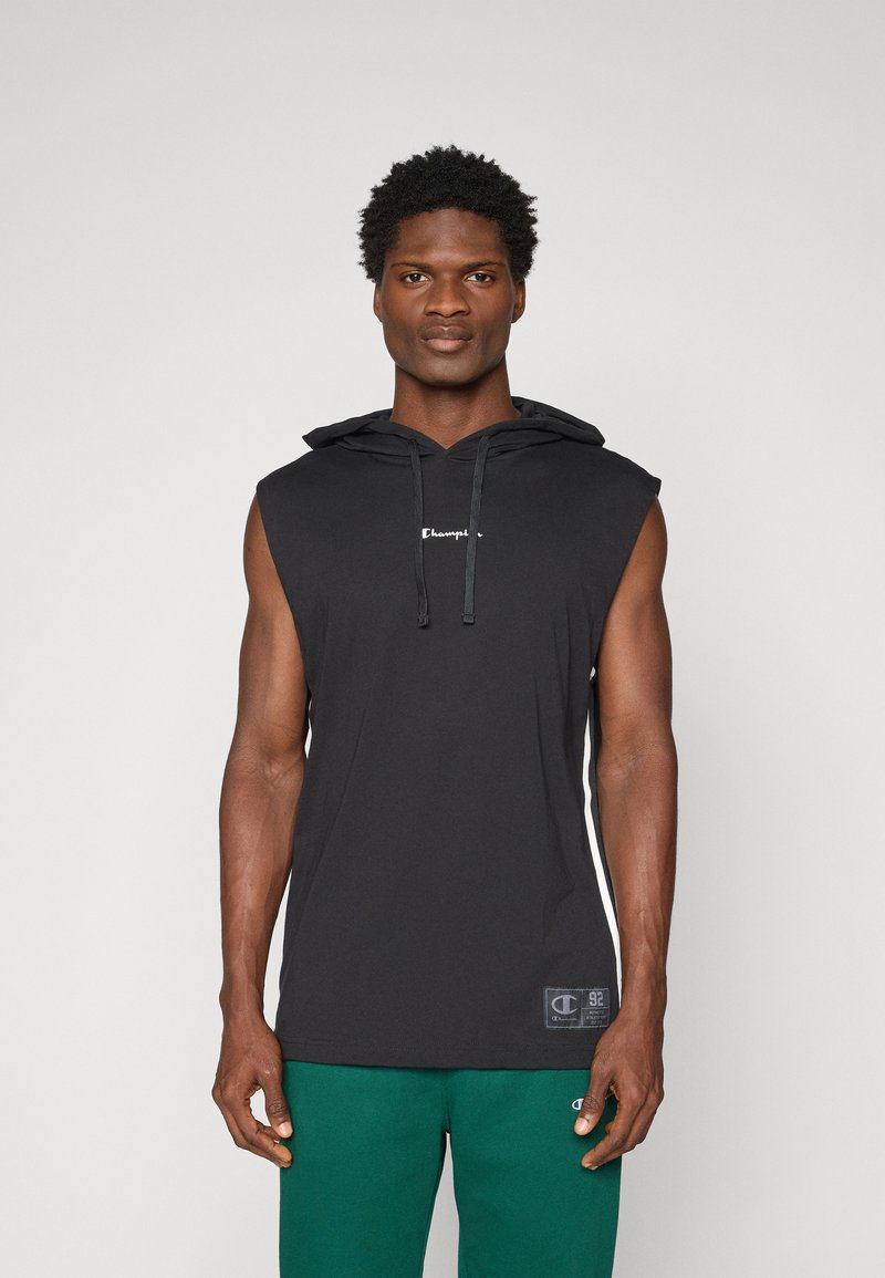 Uomo che indossa una felpa nera senza maniche con coulisse e pantaloni verdi, in piedi davanti a uno sfondo chiaro uniforme.