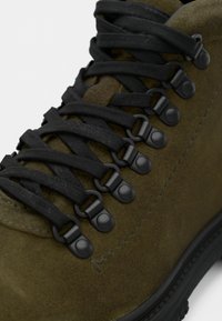 Olívovo zelená semišová turistická obuv s čiernymi kovovými D-krúžkovými očkami a hrubými čiernymi šnúrkami zviazanými do krížového vzoru.