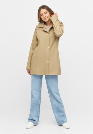 Khakifarbenes, wasserdichtes Jacket mit Kapuze, Druckknöpfen und Seitentaschen. Kombiniert mit hellblauen, weiten Jeans und weißen Sneakers.