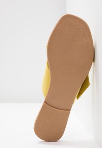 Semelle d'une chaussure plate jaune avec une semelle extérieure en caoutchouc beige texturée et un petit talon carré, positionnée debout contre un mur blanc.