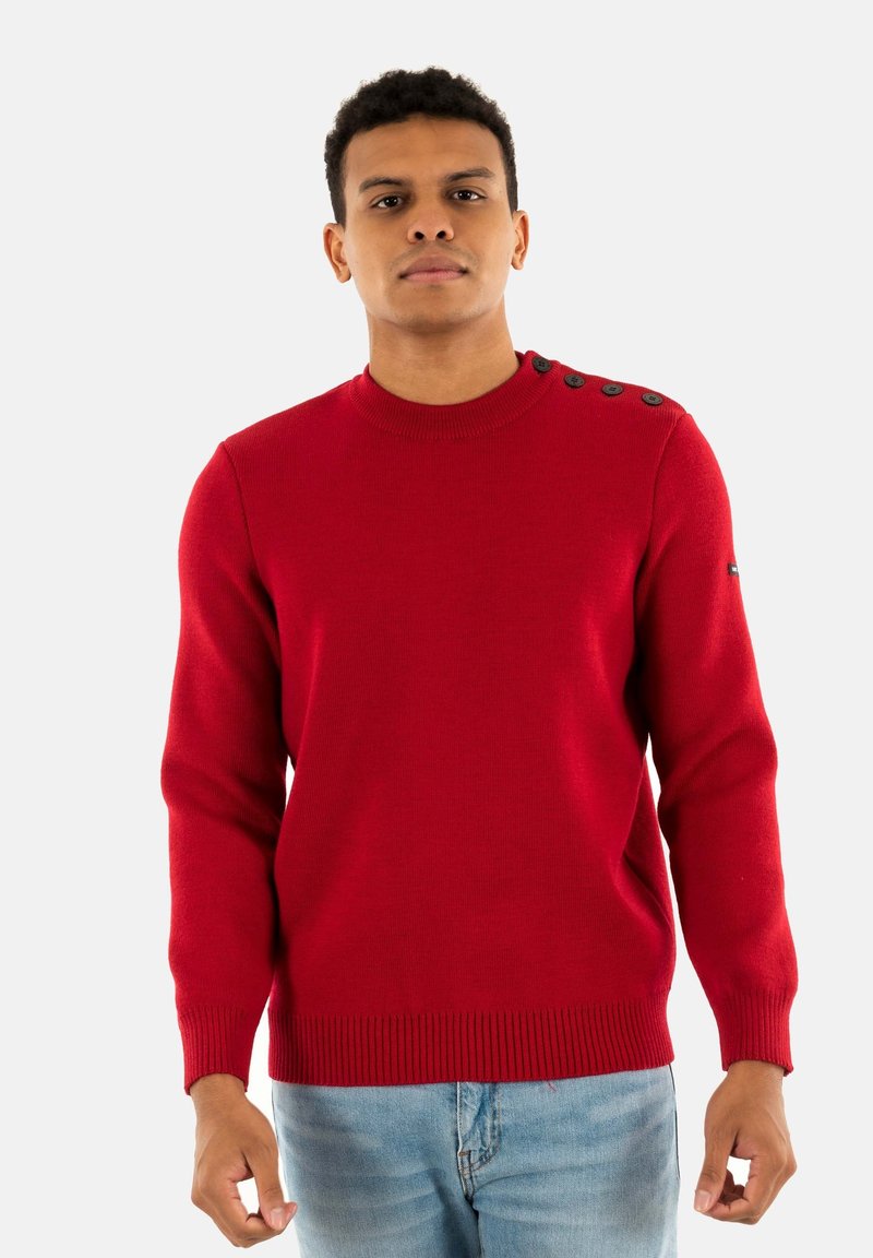 Maglione rosso a maglia con scollo rotondo, polsini e orlo a coste, e quattro bottoni decorativi sulla spalla sinistra. Indossato con jeans azzurri chiari.