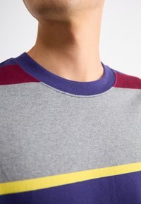 Pelēks apģērbs ar apaļu kakla izgriezumu, kas piedāvā purpursarkano un bordo krāsu blokveida detaļas, ar horizontālu dzeltenu joslu pār krūtīm. Mīksta auduma tekstūra.