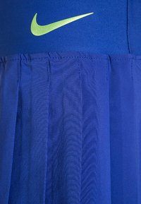 Falda deportiva azul con pliegues, que presenta un logotipo verde de Nike en la cintura. La tela parece suave con detalles de costura.