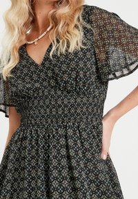 Robe noire à fleurs avec un corsage smocké, des manches flottantes en voile et des motifs blancs et verts complexes.