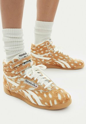 Wysokie trampki Reebok z brązowo-białym wzorem cętkowanej sierści, białymi sznurówkami i paskami, noszone z białymi, prążkowanymi skarpetami na jasnym tle.