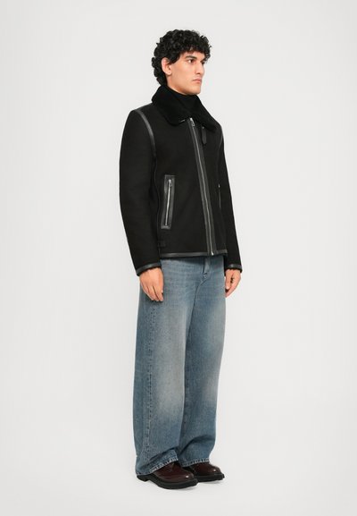 Man som står och bär en svart zip-up jacka med shearlingkrage, vida bleka blå jeans och mörkbruna skor mot en vit bakgrund.