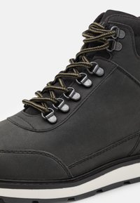 Botte de randonnée montante noire avec œillets métalliques et lacets accentués de beige, dotée d'une semelle épaisse blanche et noire crantée.