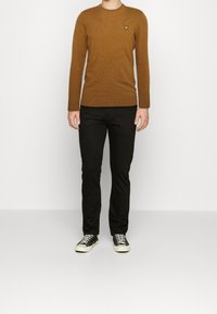 Brązowy dziergany sweter z okrągłym dekoltem i żółtym logo na piersi, zestawiony z czarnymi, dopasowanymi spodniami i czarnymi trampkami.