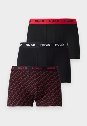Joven de pie descalzo vistiendo bóxers negros y rojos con patrón y la cintura con el logo "HUGO" sobre un fondo llano y claro.