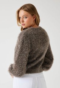 Maglione marrone fluffy con vestibilità ampia, colletto rotondo e maniche lunghe. Presenta una texture morbida e sottili accenti lucidi. Indossato con pantaloni bianchi.
