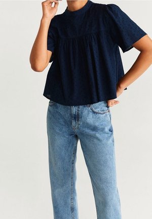 Personne portant un chemisier ample bleu marine à manches courtes avec une texture subtile à points et un jean en denim bleu clair taille haute, posant avec la main près du visage.