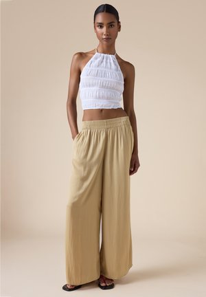 Donna che indossa un crop top bianco arricciato con collo halter e pantaloni beige larghi e ampi, in piedi davanti a uno sfondo beige uniforme.