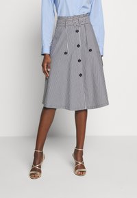 Pruhovaná sukně ke kolenům s opaskem, dvěma předními kapsami a černými knoflíky; vyrobena z lehké tkaniny v bílé a námořnické modré barvě.