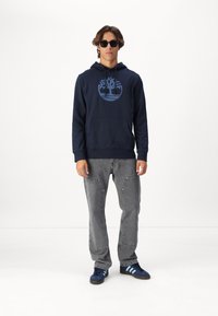 Haut à capuche bleu marine avec un motif d'arbre, poche avant, jeans gris avec des coutures décoratives, et baskets sombres avec des rayures bleues. Lunettes de soleil portées.