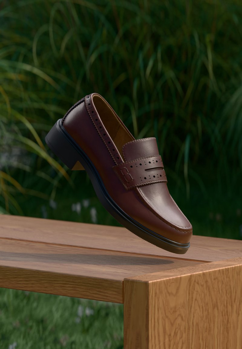 Mocassin en cuir marron avec des détails perforés, suspendu au-dessus d'une surface en bois sur un fond vert flou.