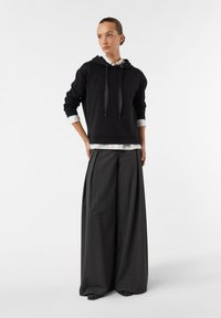 Czarna bluza z kapturem ze ściągaczami, nałożona na białą koszulę. W połączeniu z szerokimi szarymi spodniami. Gładka struktura tkaniny.