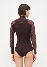 Costum de neopren lung, de culoare maro, cu mâneci din plasă cu imprimeu floral. Material texturat pe corp, cu logo-ul Roxy pe gulerul din spate. Design ajustat.