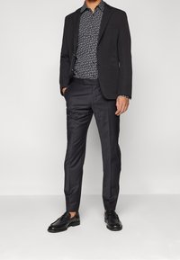 Blazer negro sobre una camisa estampada en blanco y negro, combinada con pantalones oscuros y zapatos de cuero negro. Texturas suaves y ajuste a medida.