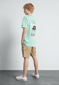 Mintgrønn bomull T-skjorte med grafisk trykk av joggesko på ryggen. Kombinert med khaki cargo shorts og hvite joggesko med røde detaljer.