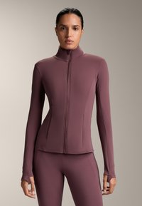 Långärmad, figurnära jacka i djupt mauve tyg med hög krage och framtida dragkedja. Har tumhål vid ärmsluten och en mjuk textur.