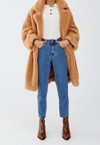 Manteau en fausse fourrure beige, haut blanc côtelé avec boutons, jean bleu taille haute, et bottes à talons imprimé serpent marron.
