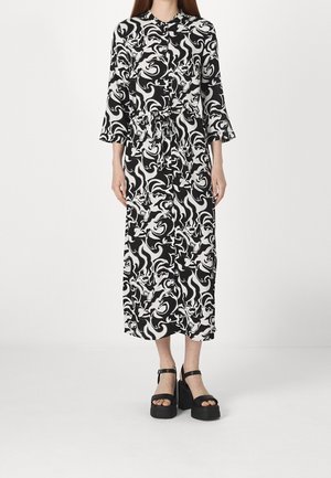 Femme portant une robe midi à manches longues avec un motif abstrait noir et blanc, fermeture boutonnée devant, et des sandales plateformes noires.