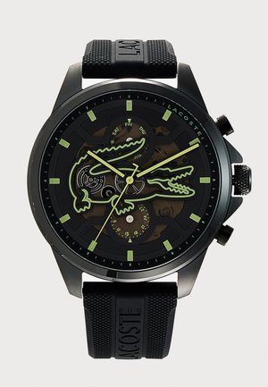 Reloj de pulsera Lacoste negro con marcadores de hora y manecillas luminiscentes verdes, con un gran contorno de cocodrilo en la esfera y una correa de goma.