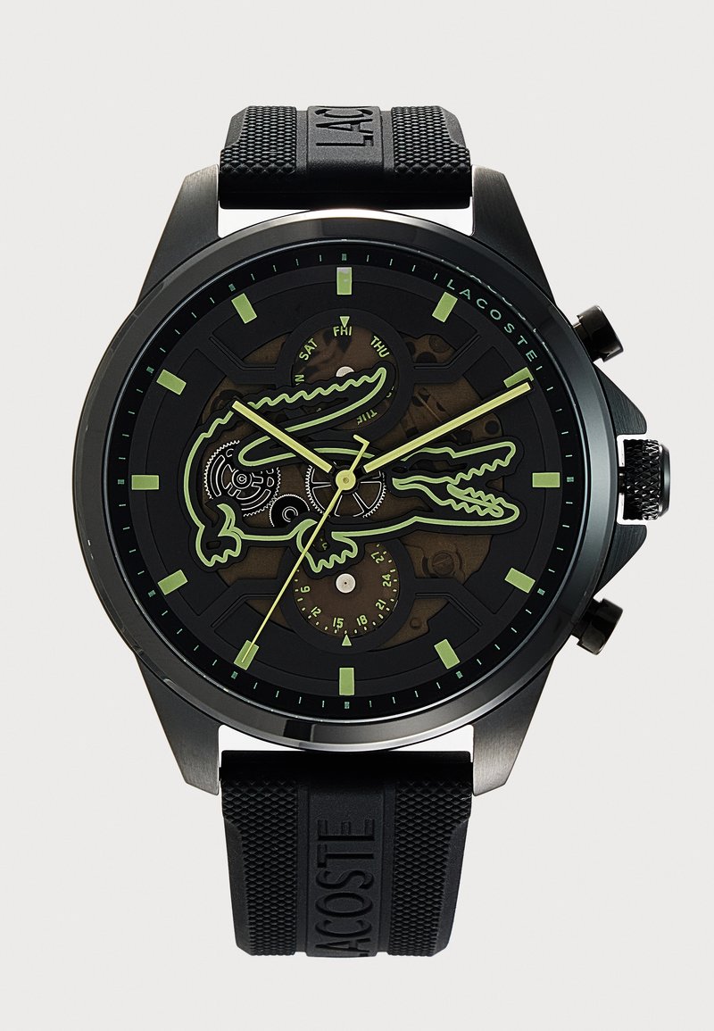 Montre bracelet Lacoste noire avec des index et des aiguilles luminescents verts, avec un grand contour de crocodile sur le cadran et un bracelet en caoutchouc.