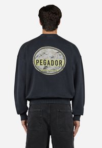 Marineblå sweatshirt med et stort cirkulært "PEGADOR" grafik på ryggen, ribbede manchetter og kant, parret med mørke jeans.