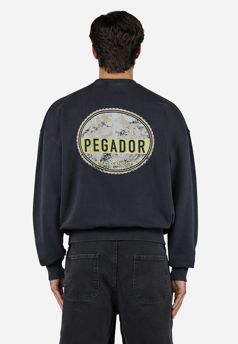 Marineblå sweatshirt med et stort cirkulært "PEGADOR" grafik på ryggen, ribbede manchetter og kant, parret med mørke jeans.
