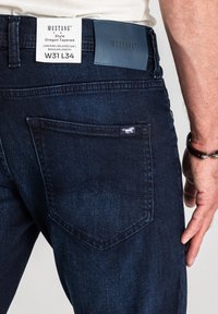 Mężczyzna noszący ciemnoniebieskie spodnie Mustang Oregon Tapered z metką pokazującą rozmiar W31 L34 na tylniej kieszeni i w pasie.