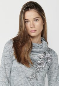 Pull encolure cheminée gris avec une texture chinée, présentant une broderie florale intricatement réalisée en blanc et gris sur le côté gauche.