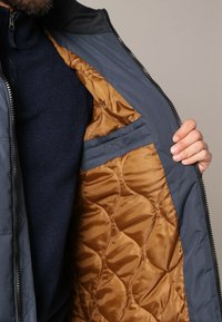 Veste bleu marine avec un doublure matelassée marron. Dotée d'un col haut et d'une fermeture éclair, elle met en valeur un tissu texturé et une poche intérieure.