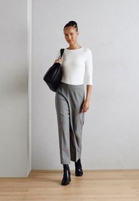 Modell iklädd en figurnära vit långärmad topp och grå byxor med hög midja, tillsammans med svarta ankelstövlar och hållande en svart handväska.
