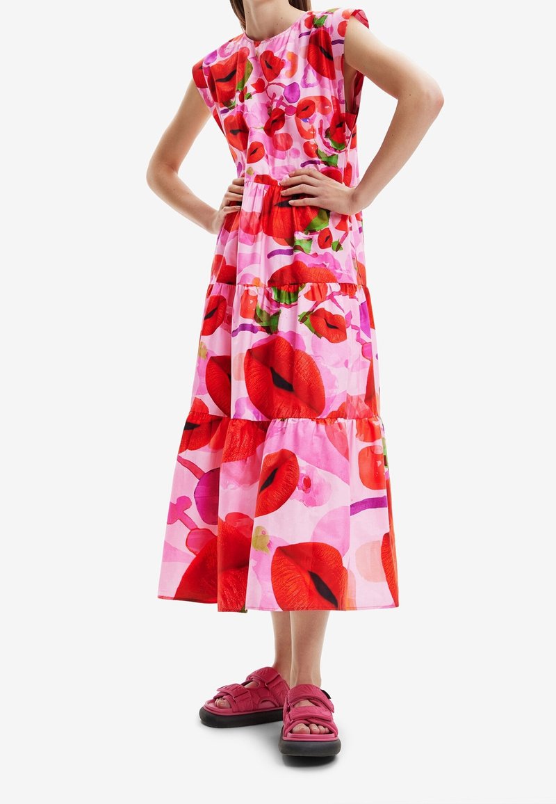 Maxi šaty z růžové látky s živým červeným motivem rtů a květinovými potisky; dvouvrstvý design s krátkými rukávy, doplněné o růžové sandály.