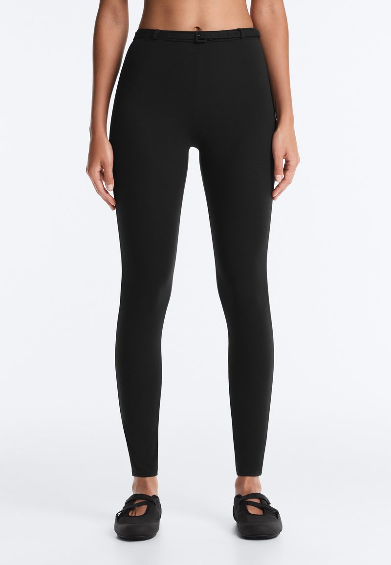 Femme portant un pantalon noir slim taille haute avec une fine ceinture noire et des chaussures plates noires à bout fermé, debout devant un fond uni.