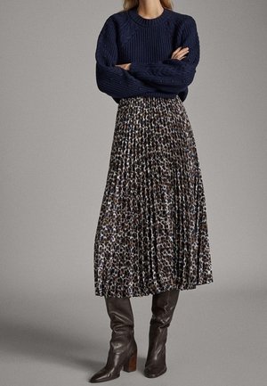 Vrouw die een marineblauwe kabelgebreide trui draagt, een geplooide midi rok met luipaardprint en donkerbruine leren kniehoge laarzen, staand met gekruiste armen.