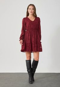 Bordowa sukienka z długimi rękawami, dekoltem w kształcie litery V i marszczonym w talii pasem. Posiada warstwowe falbany na dole oraz kwiatowy wzór na materiale.