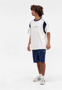 Jauns vīrietis ar cirtainiem matiem, tērpies baltā un tumši zilā oversize T-kreklā, tumši zilās šortos, baltās zeķēs un baltās kedās ar zaļām svītrām, stāv.