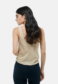 Top beige sans manches avec un dos plongeant, fabriqué en tissu léger et texturé. Assorti avec un pantalon foncé et ajusté. Cheveux naturels tombant en vagues lâches.