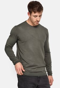 Pull en maille vert olive avec col rond et manches longues. Poignets et ourlet à côtes, mettant en valeur un design texturé et ajusté.